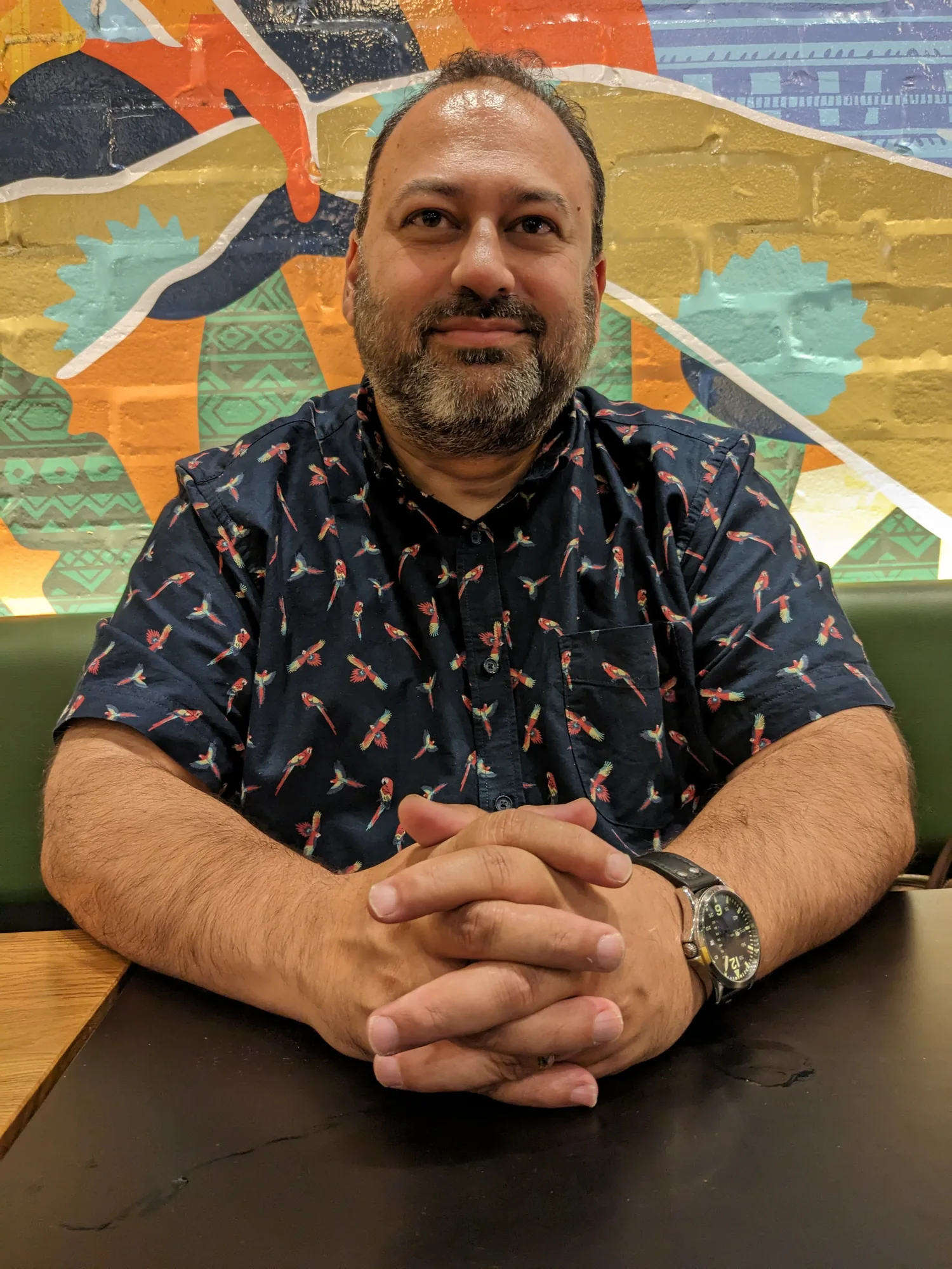 Chris Dias seated in a booth in front of a colourful mural, hands clasped, listening